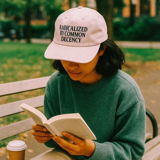 A woman sits on a park bench reading a book, wearing a light pink cap that says “Radicalized By Common Decency” in bold black text. A takeaway coffee sits beside her.