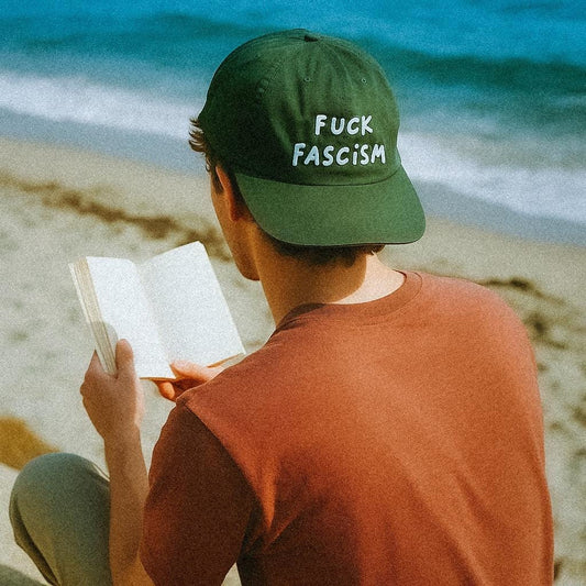 A person sits on a sandy beach reading a book, wearing a forest green cap with bold white text that reads “FUCK FASCISM” in a playful handwritten style