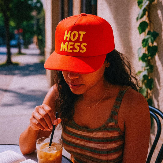 A woman in a red cap that boldly reads “HOT MESS” in yellow letters sips an iced coffee outside at a café table. She’s wearing a striped tank and minding her business—like any self-aware disaster would. Sun’s out, feelings unclear.