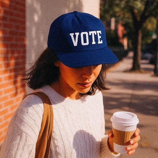 A woman in a red cap that boldly reads “HOT MESS” in yellow letters sips an iced coffee outside at a café table. She’s wearing a striped tank and minding her business—like any self-aware disaster would. Sun’s out, feelings unclear.
