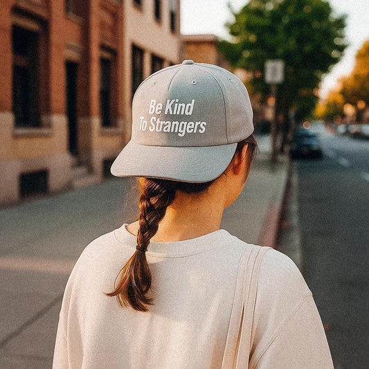 A person with a single braid walks down a quiet city street at golden hour, wearing a light grey cap made from recycled quick-dry nylon with the phrase “Be Kind To Strangers” printed in white on the back. The person is dressed casually in an off-white sweatshirt and carries a cream-colored tote bag.