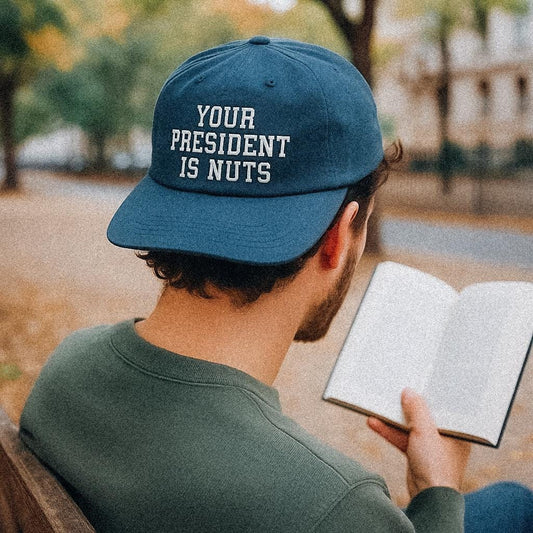 A man sits on a park bench reading a book, wearing a dark teal cap made from recycled quick-dry nylon. The cap has the phrase “YOUR PRESIDENT IS NUTS” printed in white block letters on the back. Autumn leaves scatter the path in the blurred background.