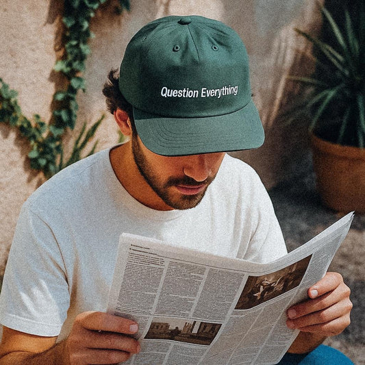 A man sits outside reading a newspaper while wearing a deep green cap made from recycled quick-dry nylon. The cap is printed with the phrase “Question Everything” in clean white text. He wears a plain white t-shirt, and potted plants with climbing vines frame the background.