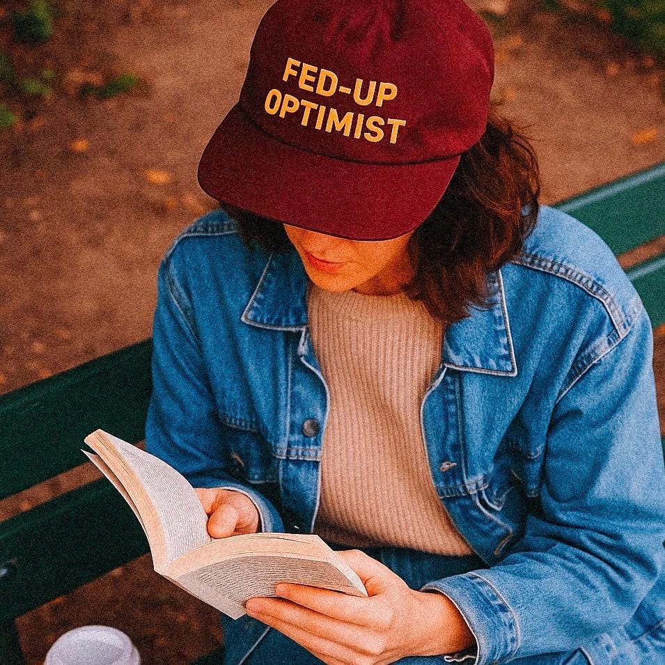 A woman reads a book while sitting on a green park bench, wearing a maroon hat that reads “FED-UP OPTIMIST” in bold yellow text. She’s dressed in a beige sweater and denim jacket, with a takeaway coffee cup beside her. The scene captures cozy autumn vibes and a hint of millennial irony.