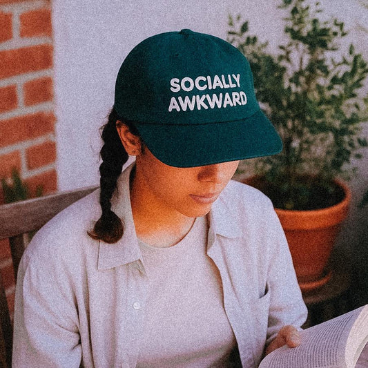 A young woman sits at a wooden outdoor table reading a book, wearing a dark green cap with the words “SOCIALLY AWKWARD” in bold white text. She’s surrounded by potted plants and dappled sunlight, perfectly embodying introvert garden chic.