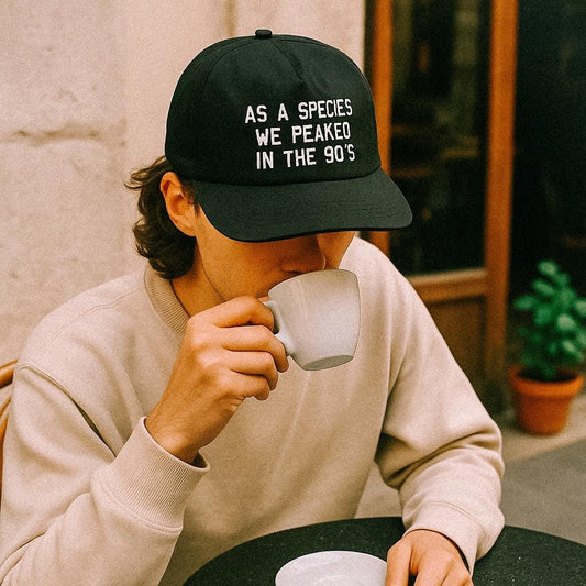 A person in a beige sweatshirt sips from a white cup at an outdoor café while wearing a black cap that reads “AS A SPECIES WE PEAKED IN THE 90’S.”