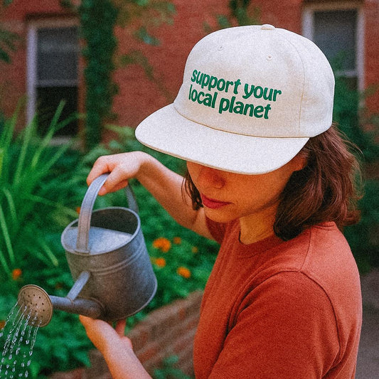 A woman waters a garden with a metal watering can, wearing a cream-colored cap that reads “support your local planet” in green lowercase letters. The mood is earthy, earnest, and eco-conscious.