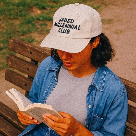 Alt text:
A woman sits on a park bench reading a book while wearing a cream-colored cap that reads “JADED MILLENNIAL CLUB” in bold black text. She’s dressed in a denim shirt over a grey tee, with sunlight casting a warm glow across the scene.