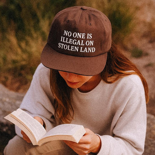 Alt text:
A woman wearing a brown recycled nylon cap that reads “NO ONE IS ILLEGAL ON STOLEN LAND” sits outdoors, reading a book. She’s dressed in a cozy, neutral-toned outfit, with soft natural lighting highlighting the thoughtful, peaceful atmosphere of the moment.