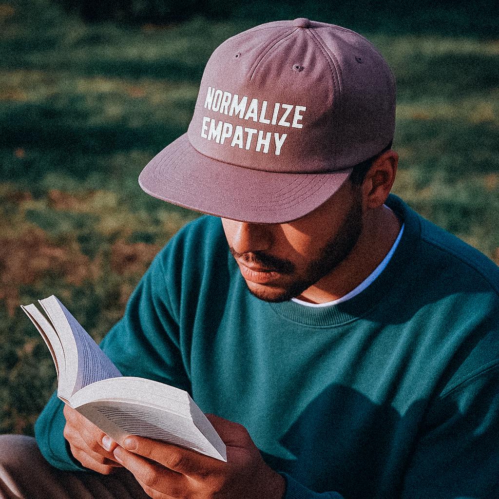 A man sits outdoors on the grass reading a book, wearing a mauve cap made from recycled quick-dry nylon. The cap features the phrase “NORMALIZE EMPATHY” printed in off-white letters. The lighting casts soft shadows across his green crewneck sweatshirt.