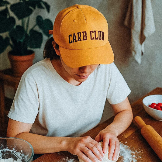A person wearing a mustard yellow cap with the words “CARB CLUB” embroidered in brown letters kneads dough on a floured wooden table. They are dressed in a plain white t-shirt, with a rolling pin, a bowl of flour, and a bowl of cherry tomatoes visible nearby. The hat is made from recycled, quick-dry nylon.